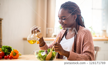 Happy young black lady preparing healthy vegetable salad in kitchen, adding olive oil and seasoning to bowl, smiling millenial african american female enjoying cooking food at home, free space 128171789