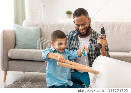 Cheerful Black Father And Son Fixing Table Using Drill And Screwdriver Doing Housework Together At Home. Household Chores For Children. Raising A Real Man Concept. Selective Focus 128171795