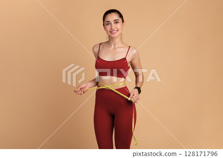 Weight Loss And Healthy Lifestyle Concept. Smiling slim woman measuring her waist with yellow tape over pastel studio background. Fit young lady in perfect shape, happy with her body 128171796