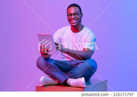 Portrait Of Cheerful Black Student Guy With Digital Tablet Sitting On Cube Under Neon Light, Smiling African Man Wearing Eyeglasses, Enjoying Modern Technologies For Education, Purple Background 128171797