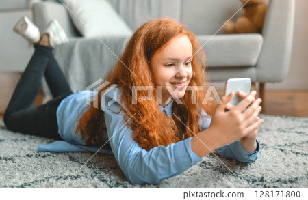 Digital Natives. Portrait of cheerful red-haired teen girl using her smartphone, chatting and texting in social media, resting and lying on the floor carpet in living room at home. Child having fun 128171800