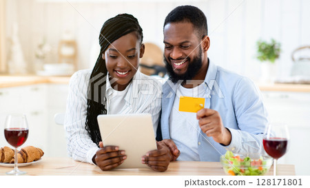 Cheerful Black Spouses Ordering Food Online With Digital Tablet And Credit Card, Sitting At Table In Kitchen, Purchasing Grocery Delivery, Enjoying Modern Technologies For Daily Life, Closeup 128171801