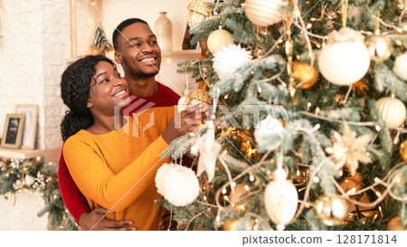 Home decor for winter holidays together at home. Millennial happy african american guy and lady decorate Christmas tree with toys, balls and garlands in living room interior, free space Home decor for winter holidays together at home. Millennial happy african american guy and lady decorate Christmas tree with toys, balls and garlands in living room interior, free space 128171814