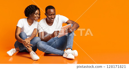 Cheerful black couple sitting together on floor and using digital tablet over orange studio background, copy space. African american happy woman and man playing games on digital pad Cheerful black couple sitting together on floor and using digital tablet over orange studio background, copy space. African american happy woman and man playing games on digital pad 128171821