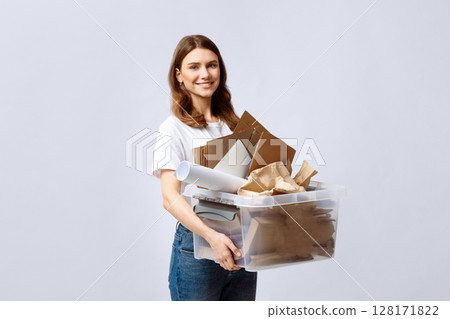 A young woman smiling while holding a transparent container filled with recyclable materials like cardboard and paper. She stands against a light background, promoting eco-friendly practices. 128171822