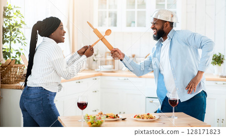 Funny Black Couple Having Fun In Kitchen, Using Kitchenware Utensils For Play Fight, Positive African American Spouses Fooling While Eating Lunch At Home, Enjoying Spending Time Together, Free Space 128171823