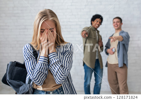 Bullying, negativity and peer problems. European and african american teens zoomers guys laugh at crying female with backpack in school or college, on white wall background, studio shot, empty space Bullying, negativity and peer problems. European and african american teens zoomers guys laugh at crying female with backpack in school or college, on white wall background, studio shot, empty space 128171824