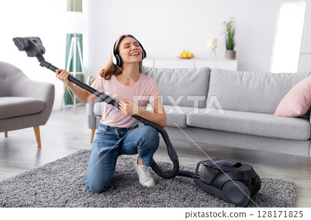 Cleaning is fun. Cheerful young housewife listening to music in headphones while vacuuming her apartment, using hoover as guitar, indoors. Playful millennial lady enjoying domestic chores Cleaning is fun. Cheerful young housewife listening to music in headphones while vacuuming her apartment, using hoover as guitar, indoors. Playful millennial lady enjoying domestic chores 128171825