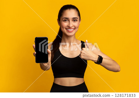 App For Healthy Lifestyle. Portrait of smiling sportswoman holding cellphone with black empty screen for mockup template and showing thumbs up sign gesture, isolated over orange studio background 128171833