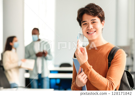 End Of The Epidemic. Smiling asian teen guy student taking off disposable protective face mask, standing in classroom at university, college or high school. Education And Healthcare Concept. End Of The Epidemic. Smiling asian teen guy student taking off disposable protective face mask, standing in classroom at university, college or high school. Education And Healthcare Concept. 128171842