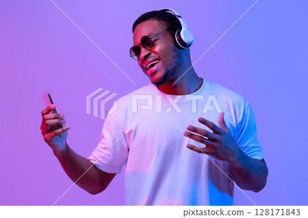 Carefree Black Guy In Wireless Headphones And Sunglasses Listening Music On Smartphone And Singing, Enjoying Favorite Songs While Standing In Neon Lighting Over Purple Studio Background, Copy Space Carefree Black Guy In Wireless Headphones And Sunglasses Listening Music On Smartphone And Singing, Enjoying Favorite Songs While Standing In Neon Lighting Over Purple Studio Background, Copy Space 128171843