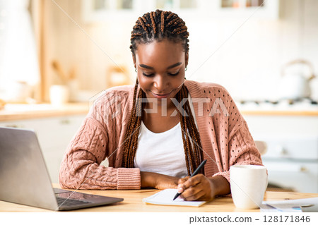 Smiling Black Student Woman Study With Laptop In Kitchen And Taking Notes, Young African American Lady Sitting At Desk At Home And Writing In Notepad, Enjoying Remote Distance Learning, Free Space Smiling Black Student Woman Study With Laptop In Kitchen And Taking Notes, Young African American Lady Sitting At Desk At Home And Writing In Notepad, Enjoying Remote Distance Learning, Free Space 128171846