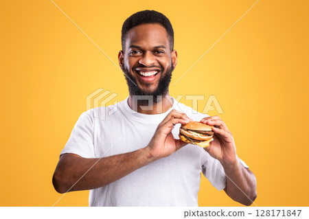 Happy black man posing holding burger smiling to camera standing over yellow background, studio shot. Junk food and cheat meal, nutrition and male diet concept. Overeating habit Happy black man posing holding burger smiling to camera standing over yellow background, studio shot. Junk food and cheat meal, nutrition and male diet concept. Overeating habit 128171847