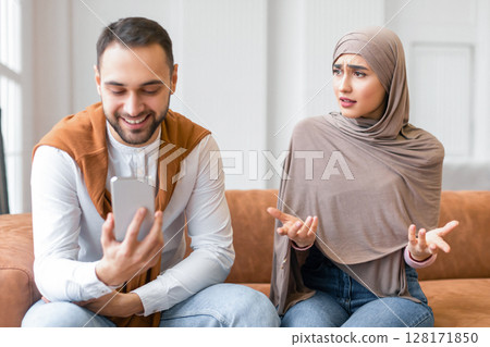 Infidelity. Displeased Muslim Wife Looking At Husband While He Texting On Smartphone Having Affair Sitting On Couch At Home. Cheating And Unfaithfulness In Marriage Concept. Relationship Problem 128171850