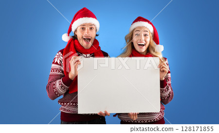Christmas ad. Excited millennial couple holding empty poster with mockup for design over blue background 128171855