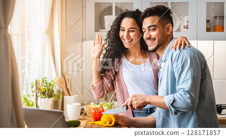 Middle Eastern Food Bloggers Couple Filming Content In Kitchen For Their Culinary Vlog, Happy Arab Man And Woman Cooking Healthy Lunch Together And Waving Hands And At Laptop Camera, Greeting Viewers Middle Eastern Food Bloggers Couple Filming Content In Kitchen For Their Culinary Vlog, Happy Arab Man And Woman Cooking Healthy Lunch Together And Waving Hands And At Laptop Camera, Greeting Viewers 128171857
