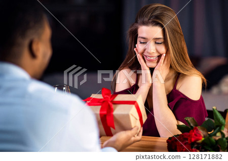 Happy International Women's Day. Loving Black Man Giving Gift To Excited Girlfriend At Romantic Dinner In Restaurant, Greeting With 8 March Holiday Or Anniversary, Multiracial Couple Dating Together 128171882