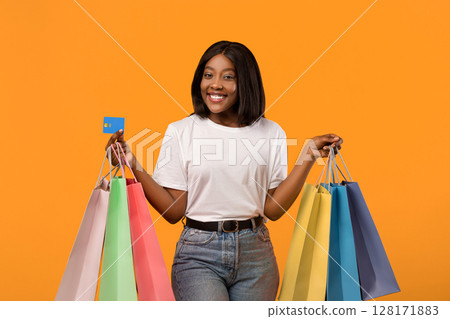 Joyful black lady with shopping bags and credit card smiling over orange studio background, empty space. African american woman shopaholic holding bunch of purchases and unlimited credit card Joyful black lady with shopping bags and credit card smiling over orange studio background, empty space. African american woman shopaholic holding bunch of purchases and unlimited credit card 128171883