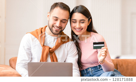 Young Arab Family Shopping Online Via Laptop Holding Credit Card Sitting On Sofa At Home. Cheerful Couple Buying Things In Internet Shop On Weekend. Discounts And Sales Concept Young Arab Family Shopping Online Via Laptop Holding Credit Card Sitting On Sofa At Home. Cheerful Couple Buying Things In Internet Shop On Weekend. Discounts And Sales Concept 128171888