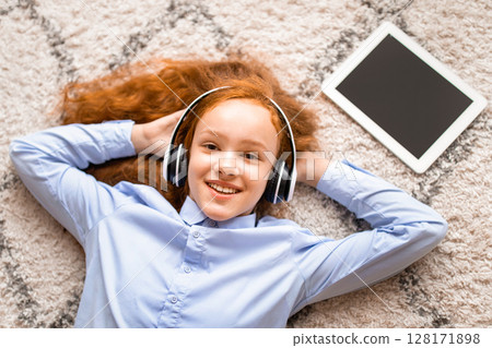 Rest And Relax. Top above high angle view of happy cheerful red-haired teenage girl listening to audio book, enjoying song wearing wireless headset, lying on floor carpet, looking at camera 128171898