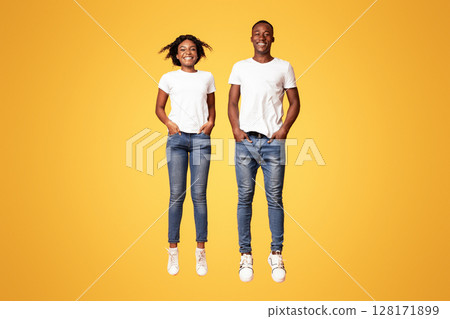 Happy black lovers young man and woman jumping up with hands in pockets, full length shot, orange studio background. Active and lifeful african american couple bouncing together 128171899