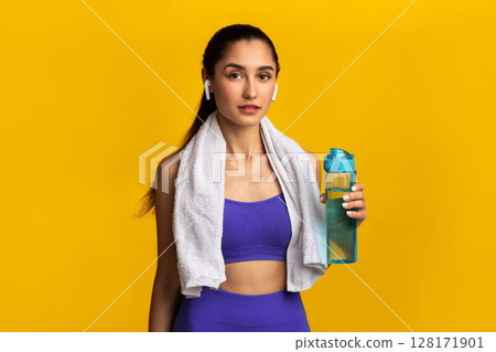 Stay Hydrated. Portrait of confident sporty young lady with white towel on neck holding bottle with fresh mineral water, wearing wireless earbuds, standing and posing isolated onorange studio wall Stay Hydrated. Portrait of confident sporty young lady with white towel on neck holding bottle with fresh mineral water, wearing wireless earbuds, standing and posing isolated onorange studio wall 128171901