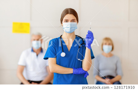 Portrait of female doctor in medical mask and uniform holding syringe with covid-19 vaccine, senior patients waiting for coronavirus immunization at clinic. Protection against virus concept Portrait of female doctor in medical mask and uniform holding syringe with covid-19 vaccine, senior patients waiting for coronavirus immunization at clinic. Protection against virus concept 128171910
