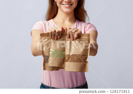 Eco-Friendly Package. Unrecognizable woman holding brown kraft paper doypack bags with weight food groceries in hands, lady buyer standing on grey background in studio, cropped image, closeup Eco-Friendly Package. Unrecognizable woman holding brown kraft paper doypack bags with weight food groceries in hands, lady buyer standing on grey background in studio, cropped image, closeup 128171911
