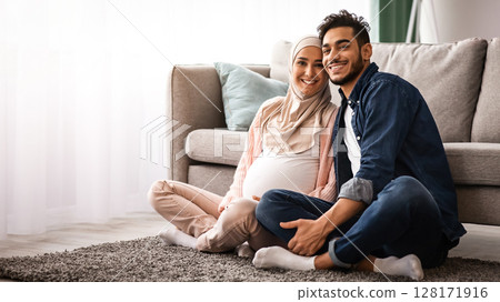 Happy Pregnant Muslim Couple Relaxing At Home Together, Sitting On Floor And Smiling At Camera. Young Arab Family With Expectant Woman In Hijab Posing To Camera In Living Room Interior, Copy Space 128171916
