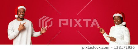 Excited african couple in santa hats pointing with fingers at free space in the middle of orange background, cheerful spouses showing empty place for your christmas offer or advertisement, panorama 128171926