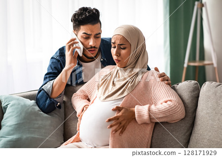 Worry Muslim Husband Calling Emergency While His Pregnant Wife Having Prenatal Contractions At Home. Islamic Expecting Mother In Hijab Suffering Painful Spasms And Spontaneous Labour, Copy Space 128171929