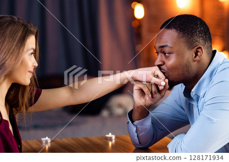 Happy Interracial Couple Flirting During Romantic Date In Restaurant, Loving African American Guy Kissing Hand Of His White Girlfriend, Multicultural Lovers Looking At Each Other, Side View 128171934