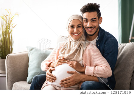 Happy religious muslim couple expecting baby posing on couch at home. Smiling expectant islamic family relaxing on sofa in living room, loving arab man hugging his pregnant wife in hijab, free space Happy religious muslim couple expecting baby posing on couch at home. Smiling expectant islamic family relaxing on sofa in living room, loving arab man hugging his pregnant wife in hijab, free space 128171939