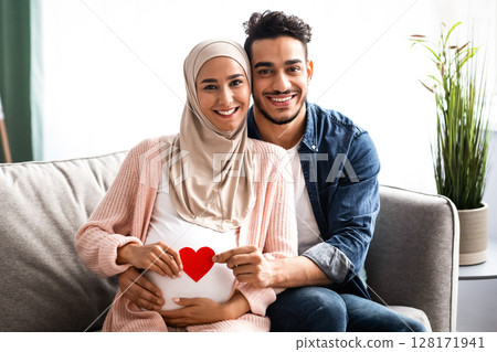 Happy Expectation. Cheerful Muslim Couple Holding Red Paper Heart Near Woman's Pregnant Belly While Posing Together On Couch At Home, Young Islamic Spouses Smiling At Camera, Free Space 128171941