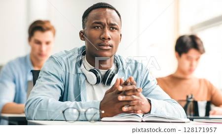 Return To Education Concept. Portrait Of Serious Focused African American Male Student Listening To Lecturer at University, Sitting At Desk In Classroom With Multiethnic Classmates 128171957