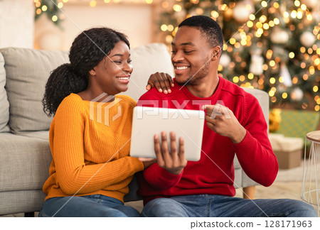 Surfing in internet and mobile app for online chat. Happy millennial african american guy points finger to tablet and shows to woman movie or sale in living room interior with Xmas tree and garlands 128171963