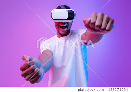 Young african american man playing video games in VR-glasses, excited black millennial guy experiencing virtual reality while standing in neon light over purple gradient background, copy space 128171964