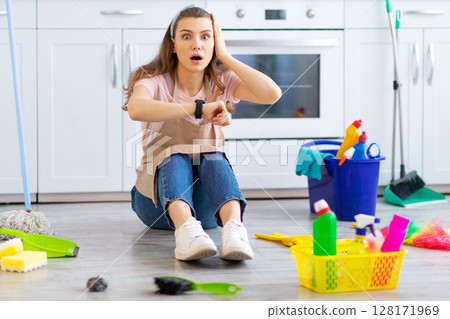 Terrified young housewife looking at her watch, being late with domestic chores on floor at kitchen. Emotional young maid missing housework deadline. Professional cleaning service concept Terrified young housewife looking at her watch, being late with domestic chores on floor at kitchen. Emotional young maid missing housework deadline. Professional cleaning service concept 128171969