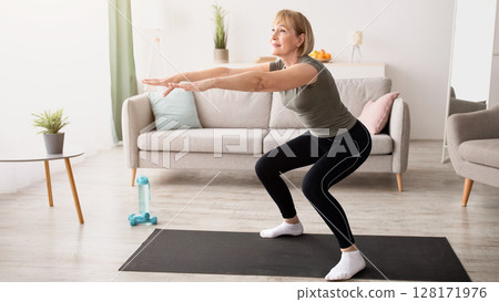 Home training concept. Active mature lady doing squats on yoga mat indoors, copy space. Sporty senior woman exercising her legs, having domestic workout during covid isolation Home training concept. Active mature lady doing squats on yoga mat indoors, copy space. Sporty senior woman exercising her legs, having domestic workout during covid isolation 128171976