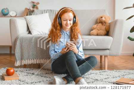 Leisure Concept. Smiling Red-Haired Schoolgirl Sitting On The Floor Leaning On The Couch At Home, Listening To Music In Wireless Headphones And Chatting On Her Cell Phone, Enjoying Free Time Leisure Concept. Smiling Red-Haired Schoolgirl Sitting On The Floor Leaning On The Couch At Home, Listening To Music In Wireless Headphones And Chatting On Her Cell Phone, Enjoying Free Time 128171977