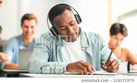 Studying And Learning Concept. Portrait of focused young black man in wireless headset sitting at table in lecture hall in highschool, writing in notebook, taking notes or examination, selective focus 128171979