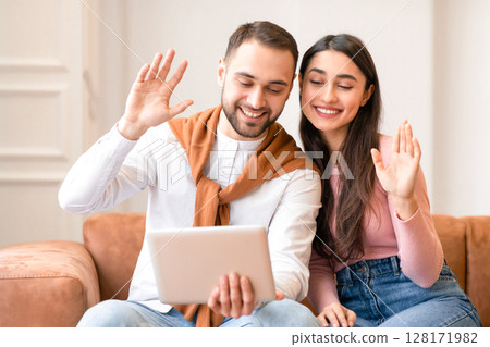 Modern Arab Couple Making Video Call Via Digital Tablet Waving Hello To Computer Webcam Sitting On Couch At Home. Middle Eastern Spouses Communicating Online With Family. Remote Communication 128171982