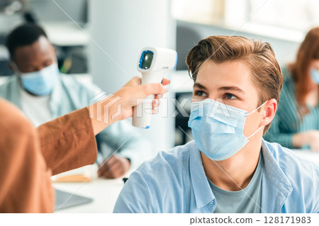 Woman using digital medical contactless infrared electronic thermometer, measuring and checking body temperature, screening male student in protective mask, preventing contagious diseases spread 128171983