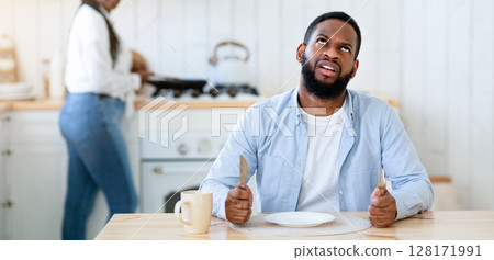 Impatient hungry black husband waiting for dinner with empty plate, bored starving african american man sitting at table and rolling eyes while his wife cooking food on background, free space 128171991