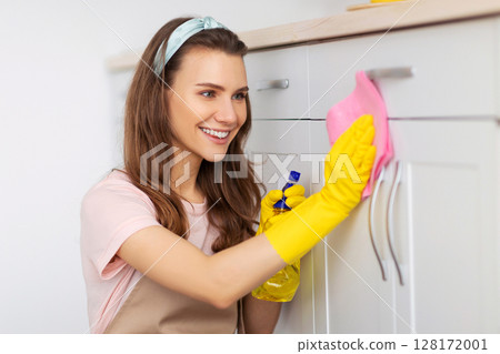 Joyful young woman cleaning kitchen cabinet with detergent and rag, keeping her home tidy. Pretty millennial housemaid doing her professional chores, performing house cleanup Joyful young woman cleaning kitchen cabinet with detergent and rag, keeping her home tidy. Pretty millennial housemaid doing her professional chores, performing house cleanup 128172001