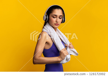 Music For Motivation. Portrait Of Determined Young Sportive Lady Wearing Wireless Headset Listening To Playlist Standing Isolated Over Orange Studio Background With White Towel Aroung Neck Music For Motivation. Portrait Of Determined Young Sportive Lady Wearing Wireless Headset Listening To Playlist Standing Isolated Over Orange Studio Background With White Towel Aroung Neck 128172002