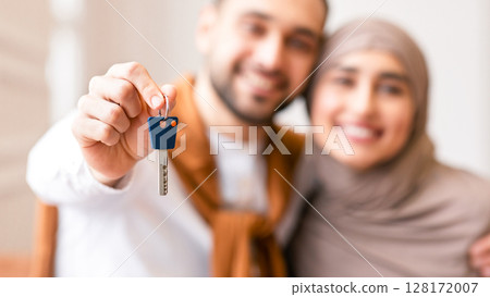 Joyful Muslim Couple Showing New House Key Standing In Their Own Home After Relocation, Focus On Key. Real Estate Ownership And Mortgage Loan, Buying New Flat Concept. Shallow Depth 128172007