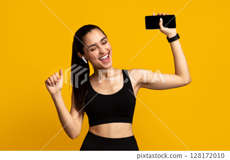 Music And Sport. Portrait Of Excited Young Fit Lady In Sportswear And Wireless Earbuds Dancing Isolated Over Orange Studio Background And Listening To Favorite Song, Holding Smartphone Music And Sport. Portrait Of Excited Young Fit Lady In Sportswear And Wireless Earbuds Dancing Isolated Over Orange Studio Background And Listening To Favorite Song, Holding Smartphone 128172010