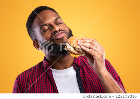 African Millennial Guy Smelling Tasty Burger Standing On Yellow Studio Background. Black Man Enjoying Eating Hamburger. Unhealthy Nutrition Habit, Overeating. Junk Food Lover Concept 128172016