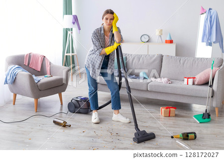 Unhappy young woman standing with vacuum cleaner in messy flat after party, blank space. Upset millennial housewife at dirty room after celebration, garbage scattered everywhere 128172027